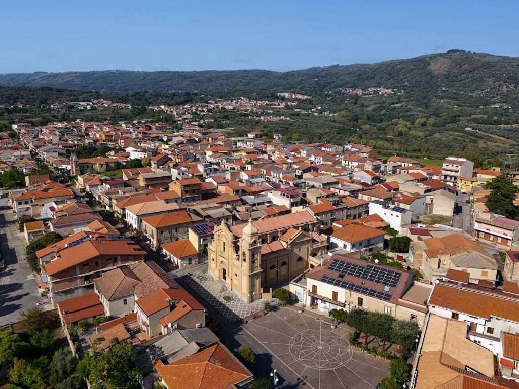 Veduta aerea di Limbadi con la piazza e la maestosa Chiesa di San Pantaleone al centro. La foto mostra i tetti del centro storico e le colline verdi sullo sfondo.